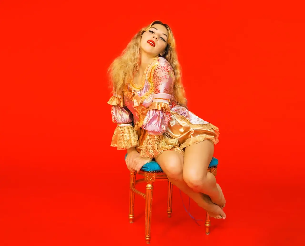 Vasilisa Forbes seduta su una sedia dorata, con costume rosa e oro davanti a uno sfondo rosso.